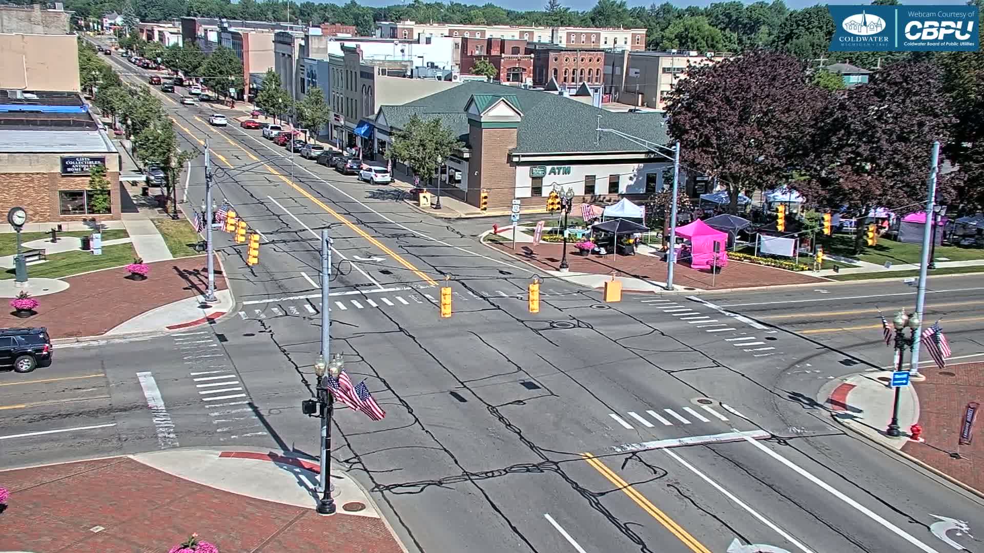 A sunny day shows a town square with a street intersection and several parked cars, alongside small vendor tents in a grassy area.