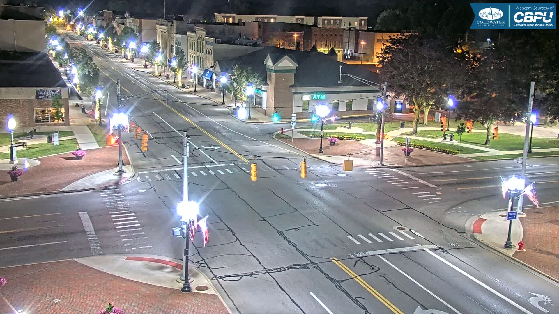 A nighttime view of a mostly empty, four-lane road intersecting with a two-lane road in a town, lined with streetlights and buildings.