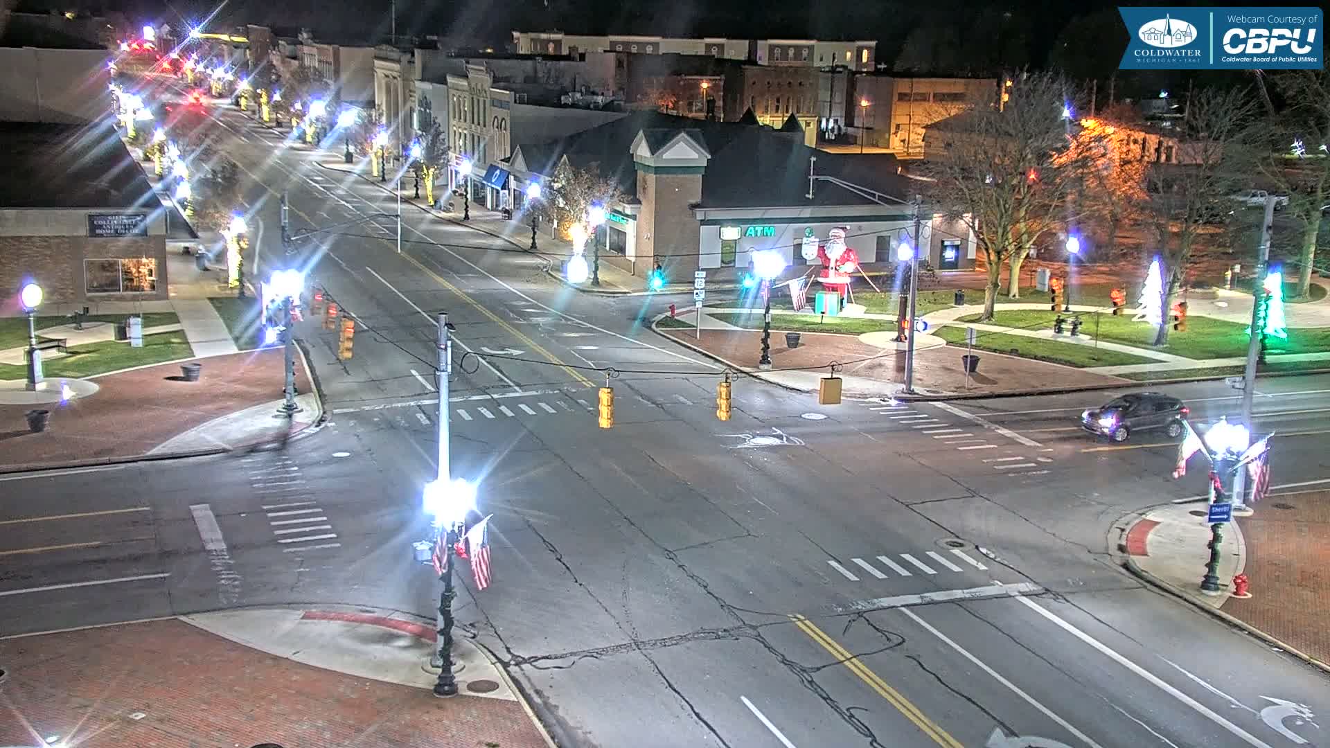 A brightly lit downtown street intersection is captured at night, adorned with festive holiday lights, illuminated trees, and a large inflatable Santa Claus, with one car driving through under clear weather conditions.