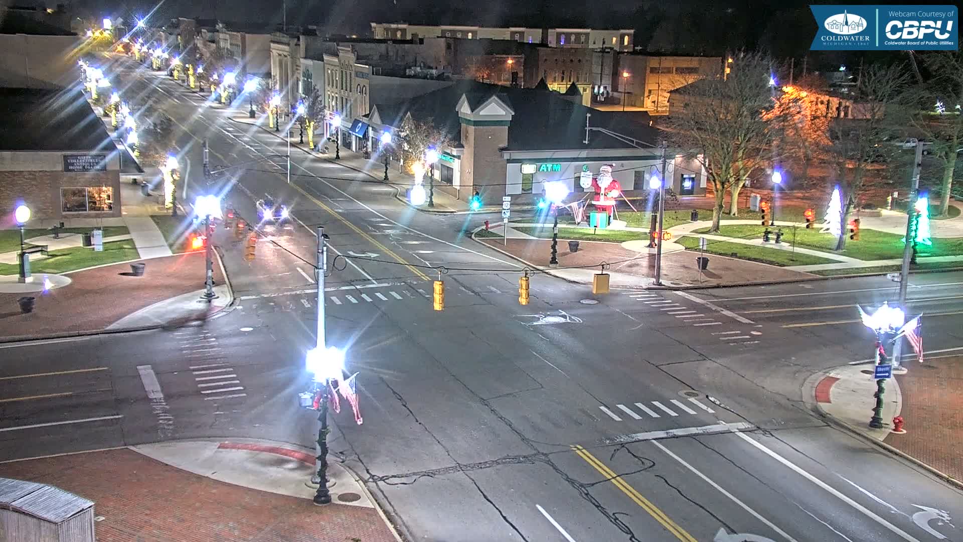 A well-lit downtown street intersection is seen at night, festive with holiday decorations including a large Santa Claus inflatable and illuminated trees, while the damp pavement reflects the bright streetlights.
