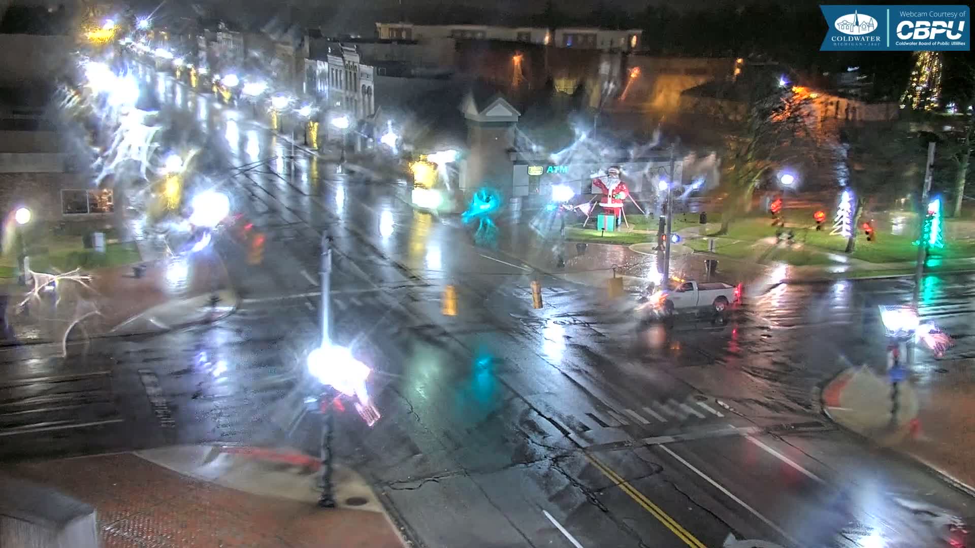 A brightly lit city street at night is slick with rain, reflecting numerous lights, and features festive Christmas decorations including a large Santa display in a median, with a white pickup truck visible at an intersection.