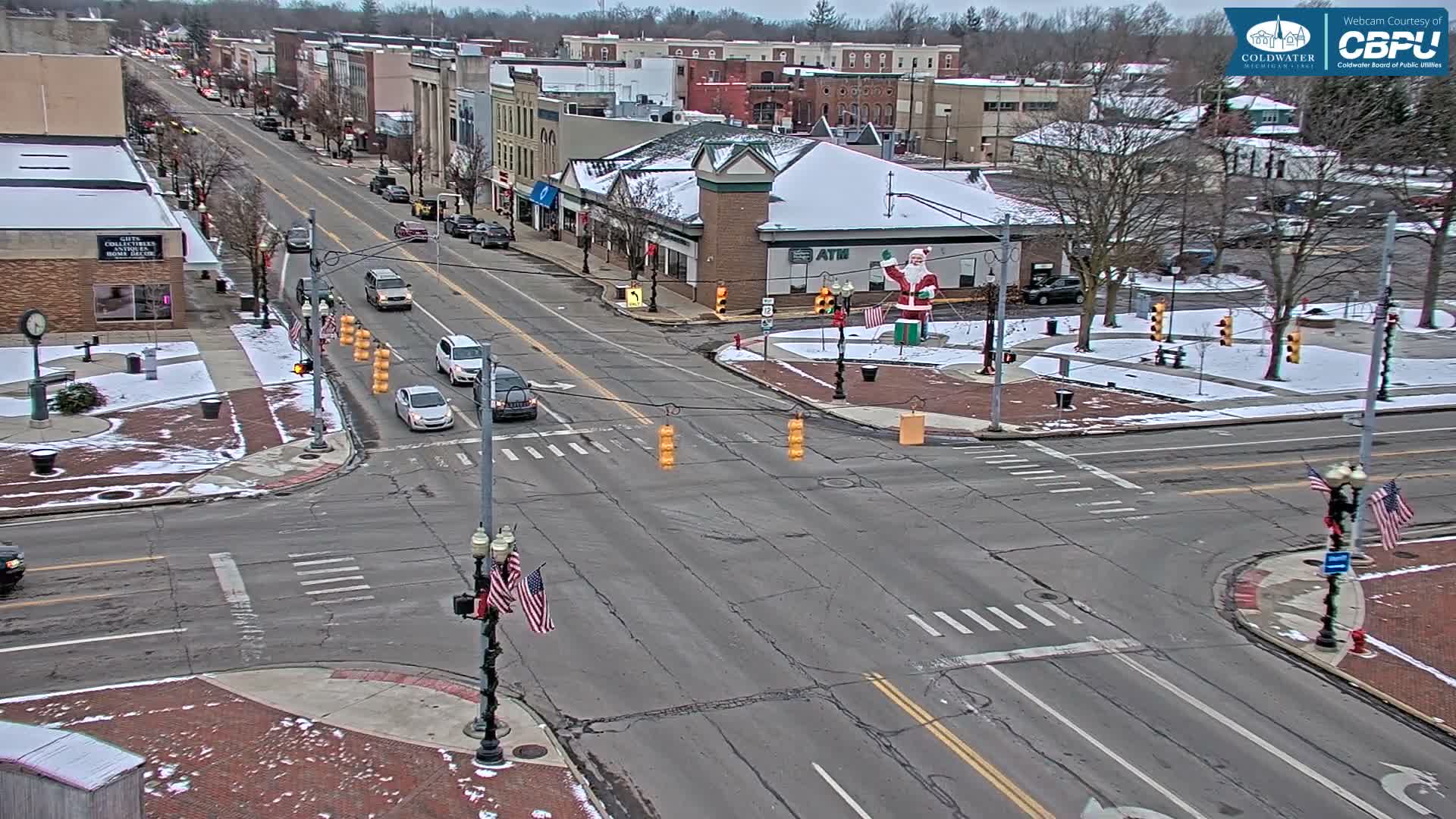 The image shows an overhead view of a bustling town intersection with cars on the road, snow covering rooftops and parts of the ground, and a large Santa Claus figure in a central median, all under an overcast winter sky.