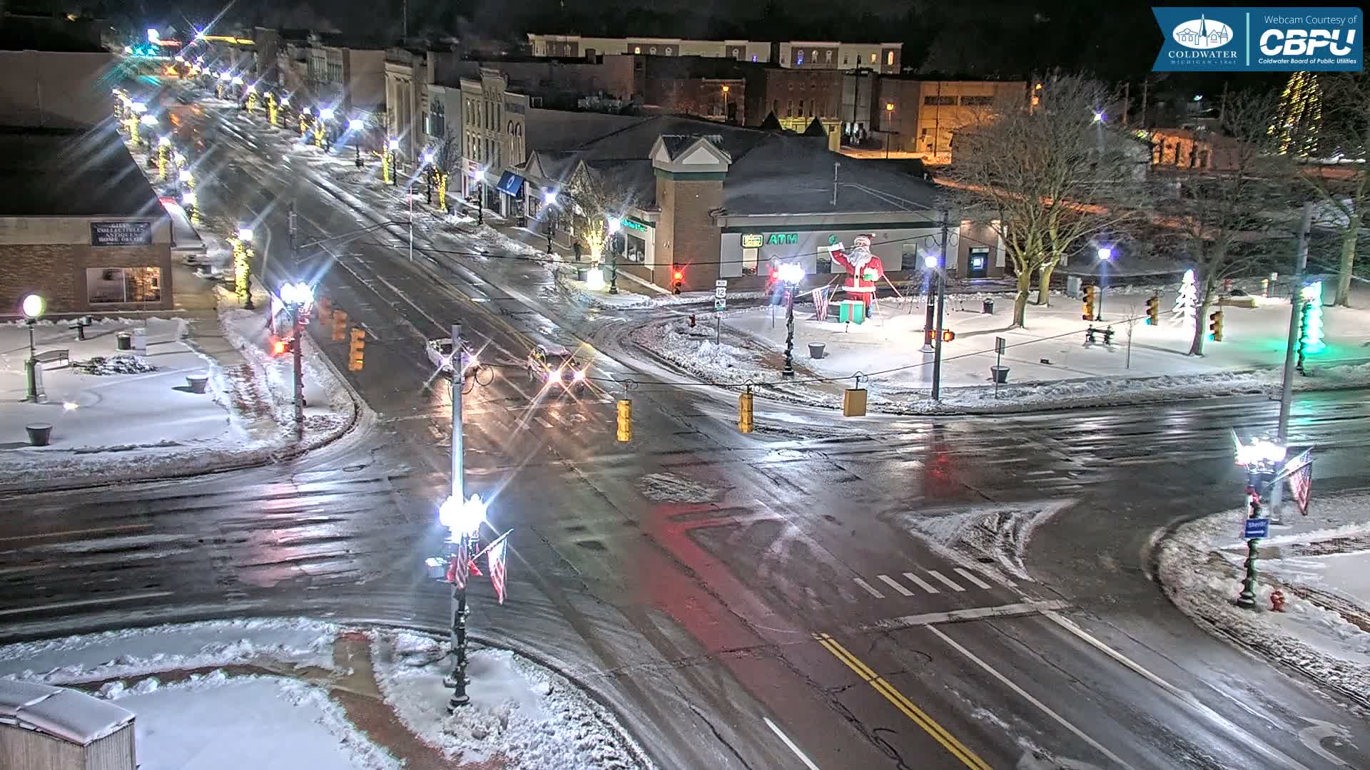 A snowy and wet urban street intersection is brightly lit at night with numerous streetlights, festive holiday decorations including a large Santa Claus, and reflections of lights on the road.