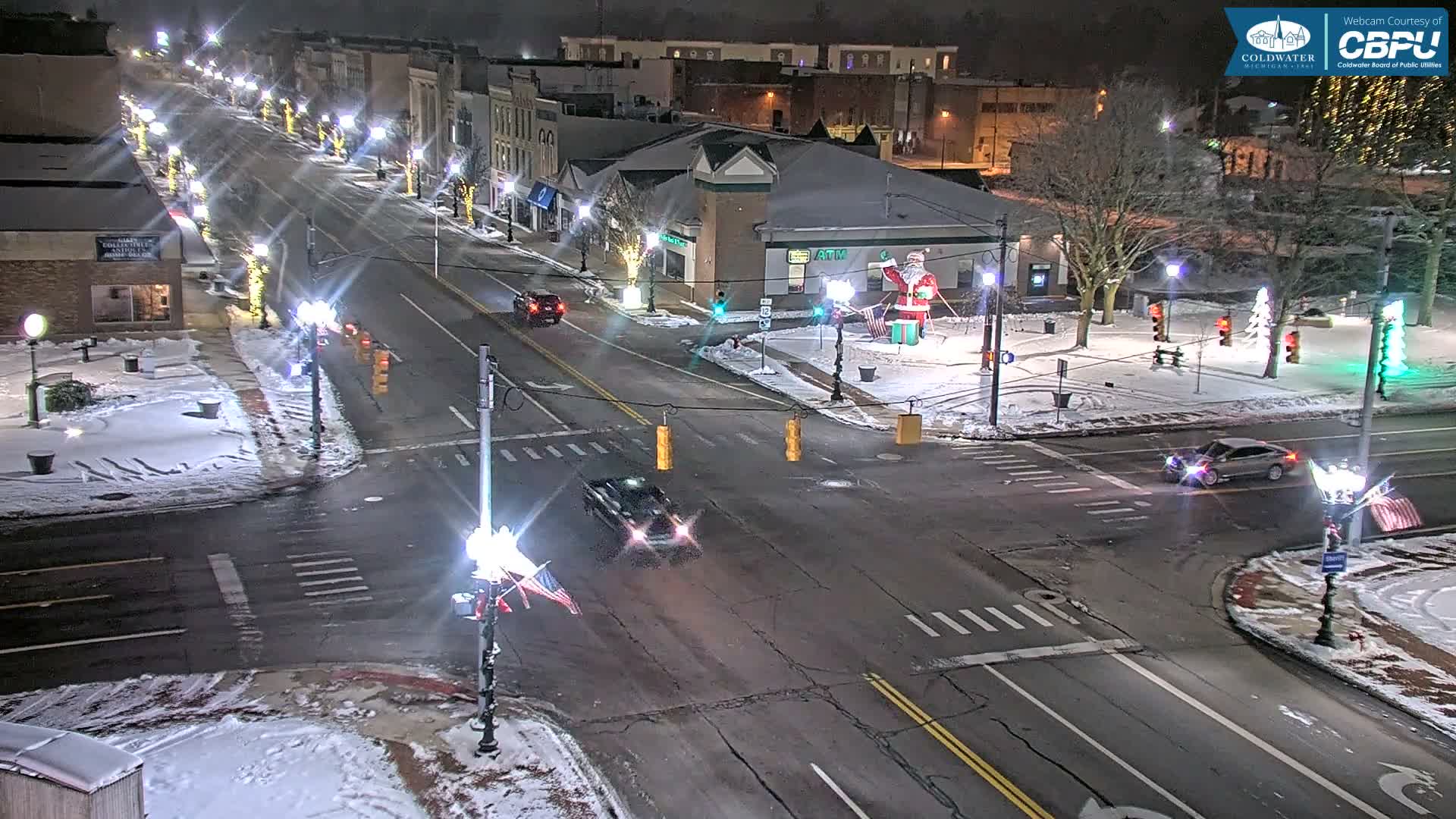 A snowy city intersection at night is brightly illuminated by streetlights and festive holiday decorations, with cars moving along the slushy roads under clear, cold conditions.