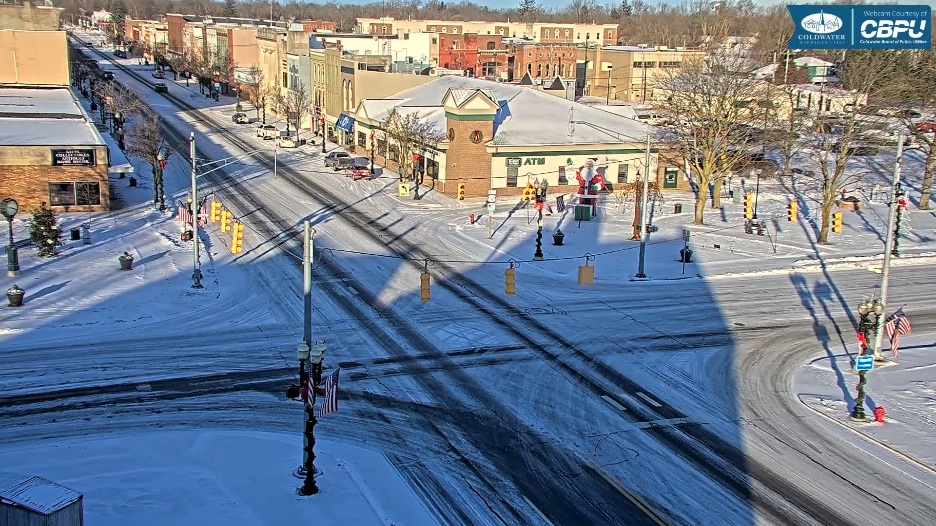 A snowy city intersection at night is brightly illuminated by streetlights and festive holiday decorations, with cars moving along the slushy roads under clear, cold conditions.