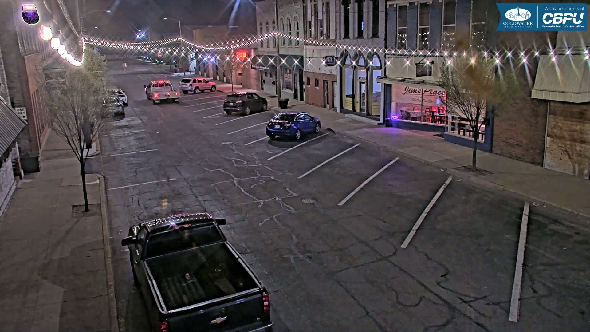 A mostly empty street at night, lined with parked cars and buildings decorated with string lights, is viewed from above.