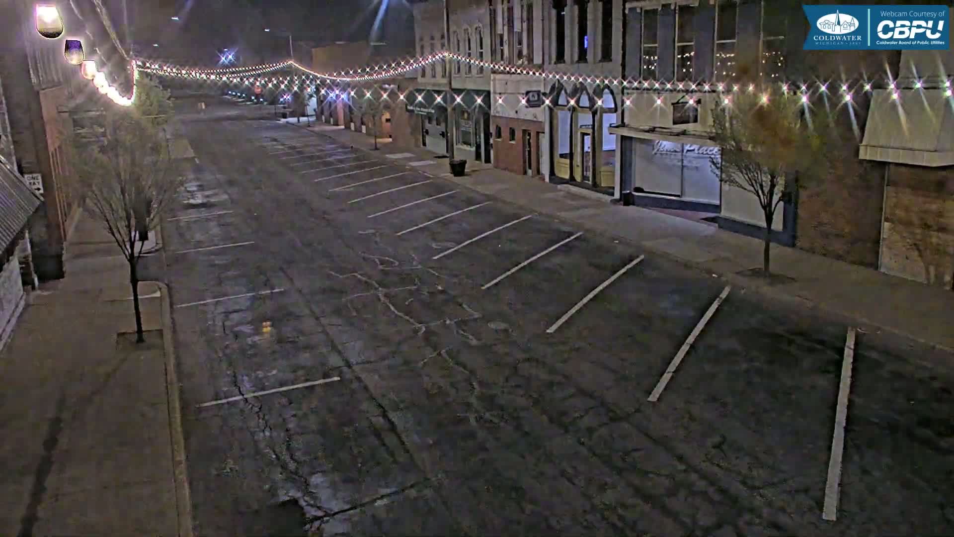 A mostly empty street at night, lined with buildings decorated with string lights, shows empty parking spaces on either side.