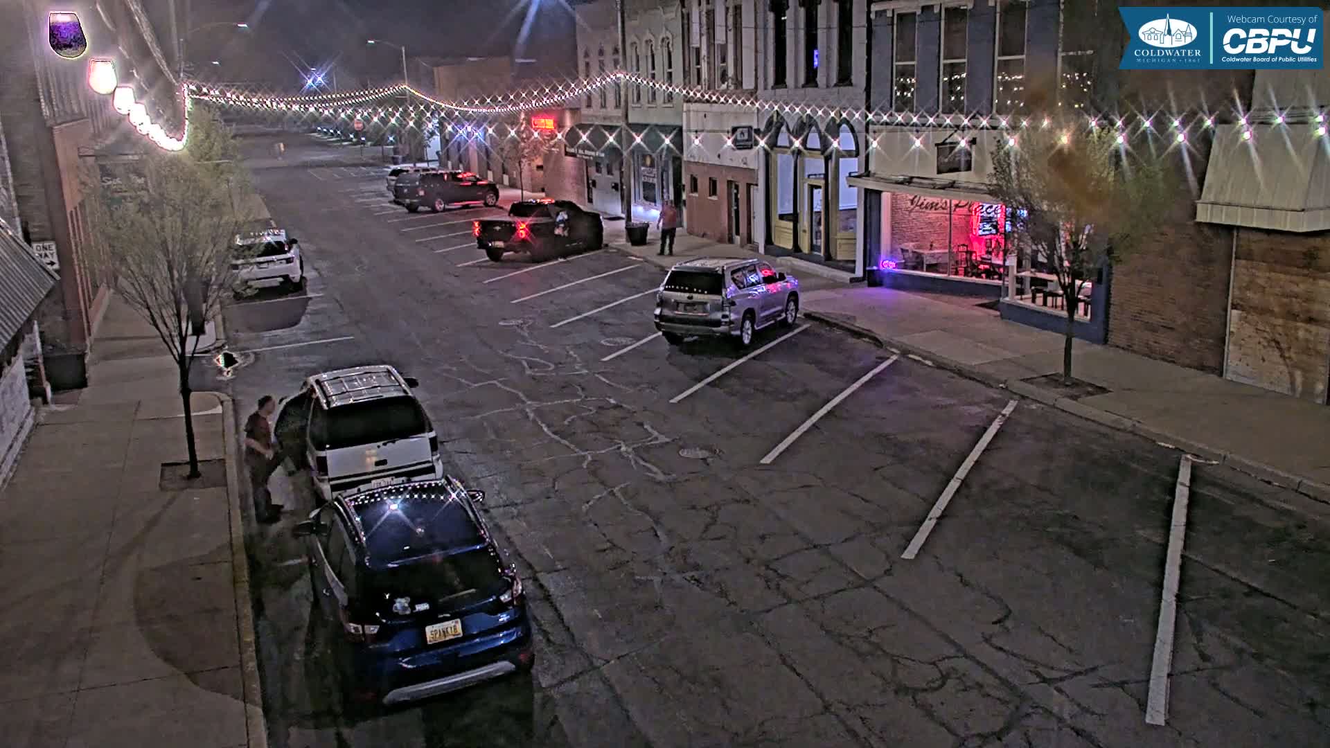 At night, several cars are parked along a street lined with brick buildings decorated with string lights.