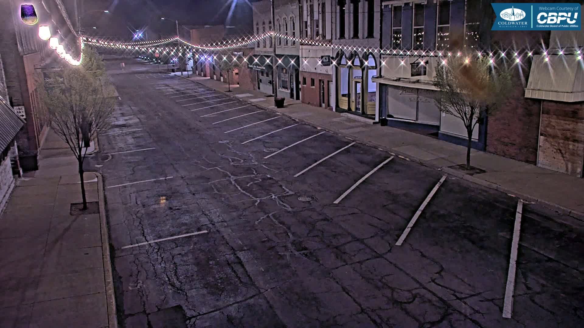 A mostly empty street at night lined with brick buildings and string lights, showing cracked pavement and some parked cars.