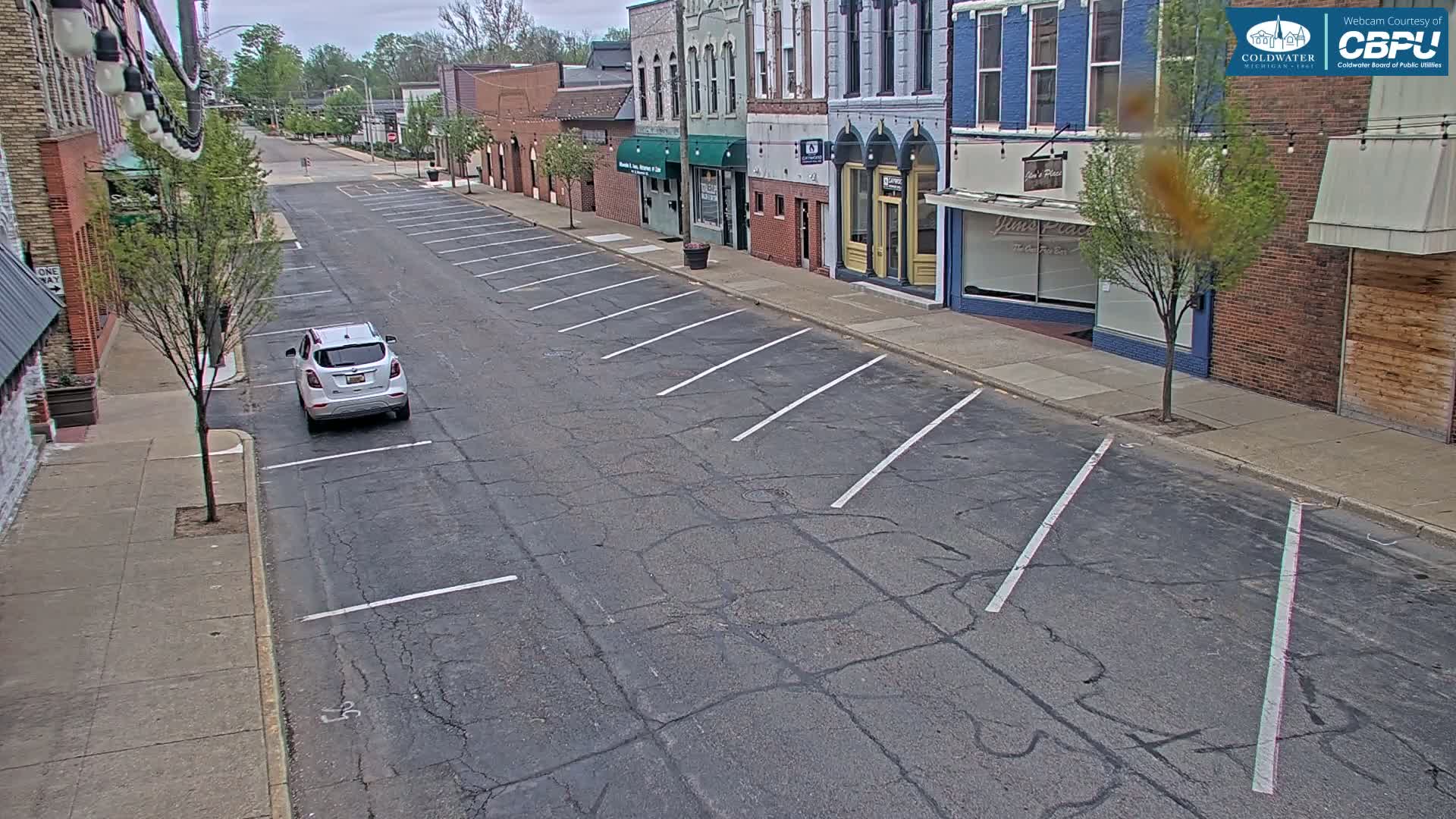 A mostly empty street with parallel parking spaces on a partly sunny day shows a white SUV parked, and several brick buildings line the street.