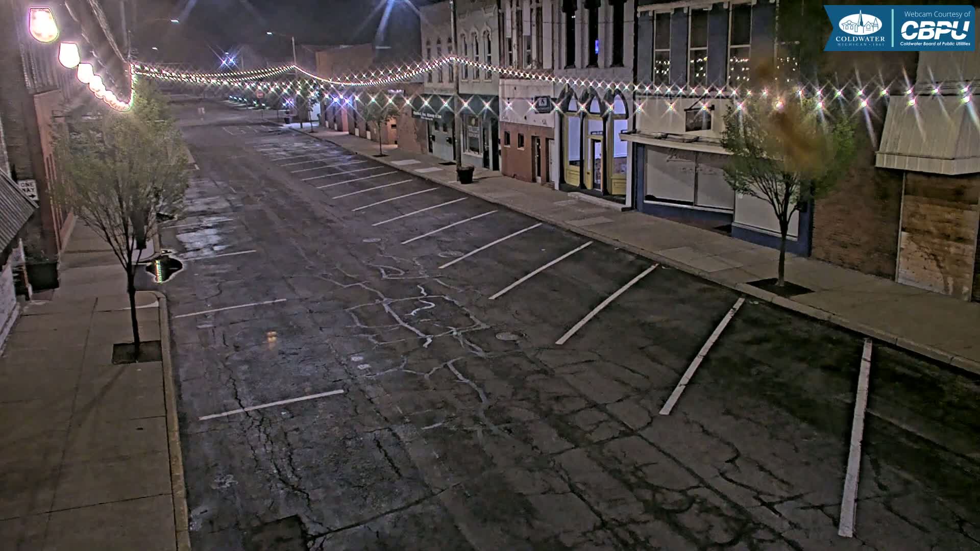 A mostly empty, nighttime street lined with buildings decorated with string lights shows a cracked asphalt parking lot and sidewalks.