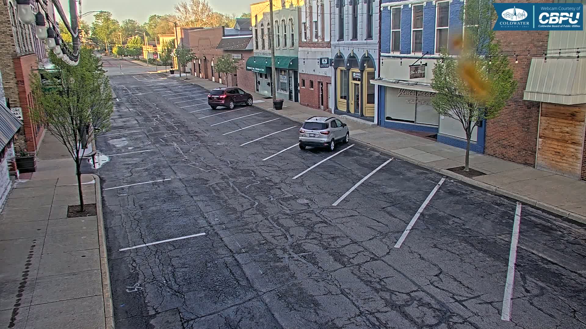 A mostly empty street lined with brick buildings on a partly sunny day, with two cars parked in a cracked asphalt parking lot.