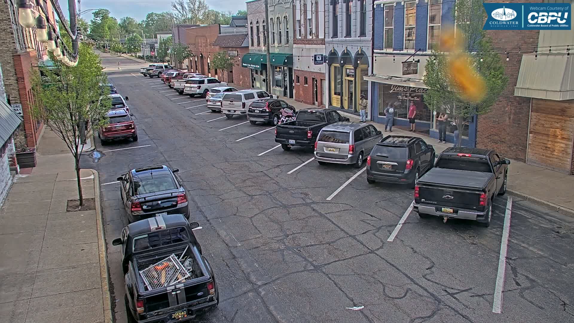 Cars are parked along a street in front of brick buildings on a partly sunny day.