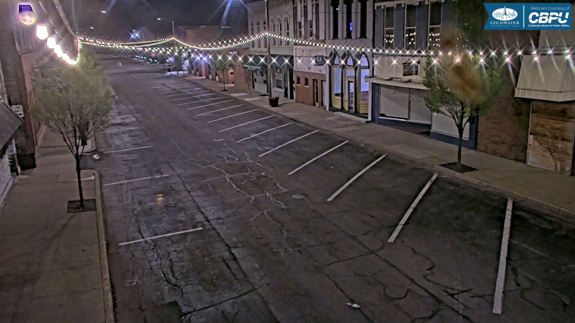 A mostly empty, nighttime street lined with buildings and parking spaces, illuminated by string lights.