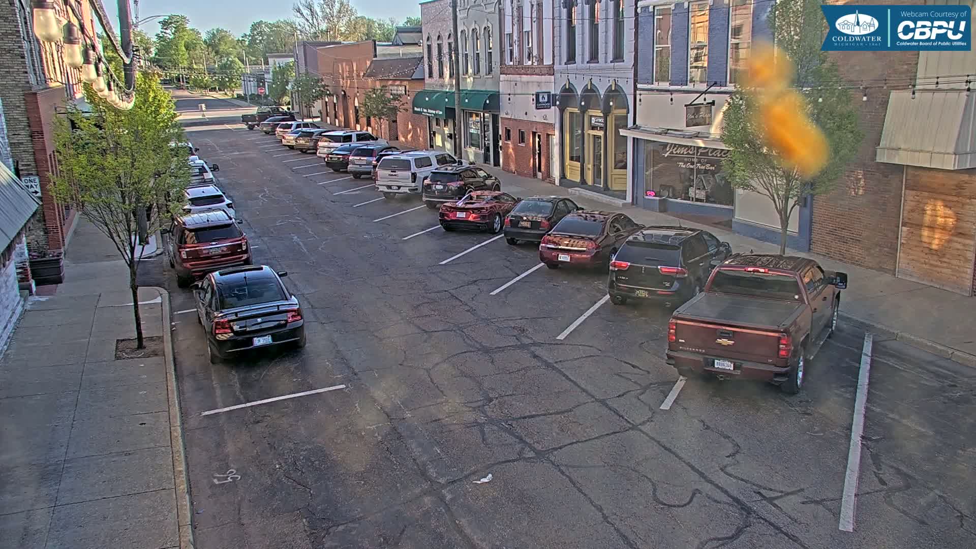 Cars are parked along a street in front of brick buildings on a sunny day.
