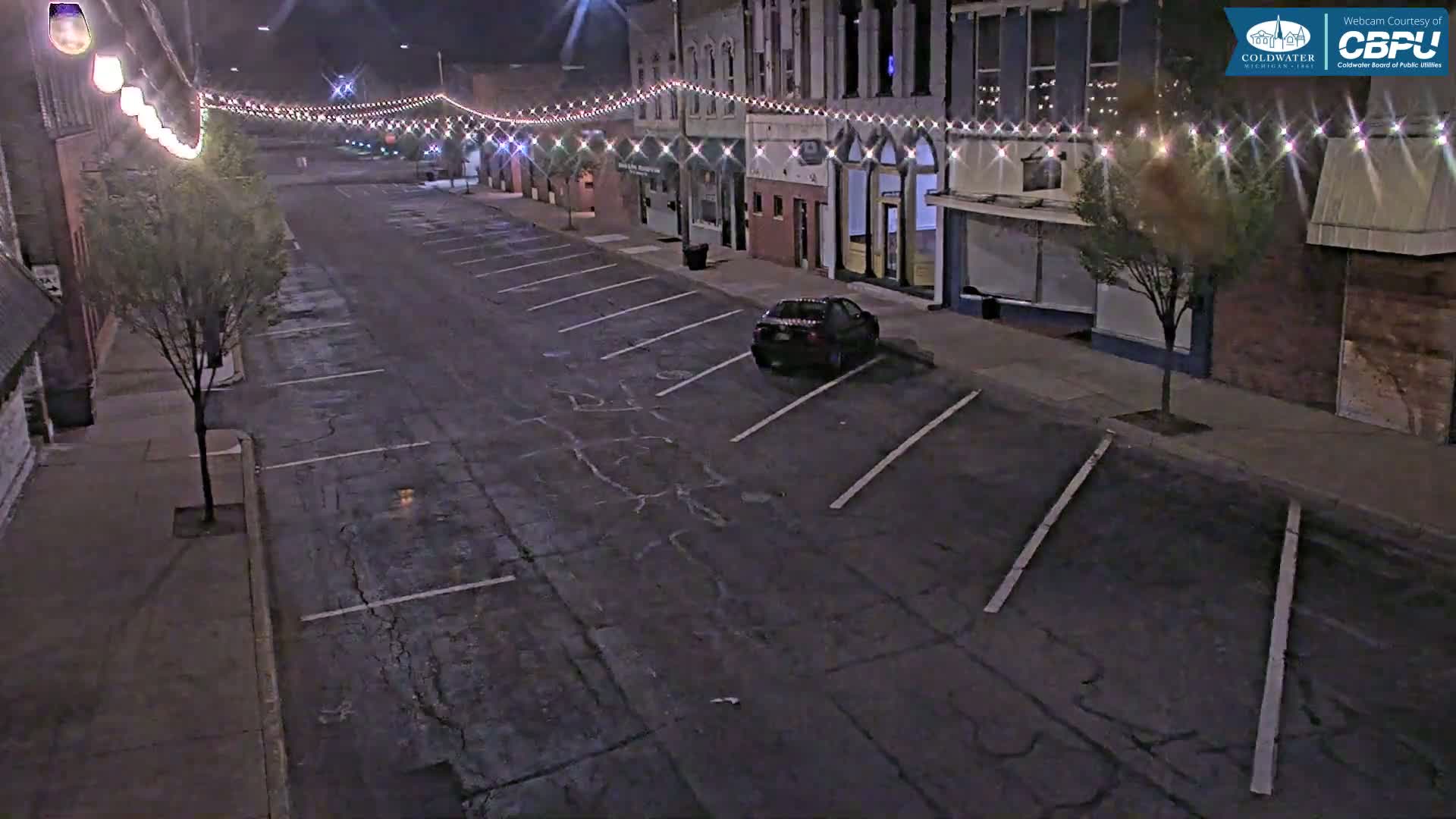 A nighttime street scene shows a mostly empty parking lot in front of brick buildings decorated with string lights, and a single car parked.