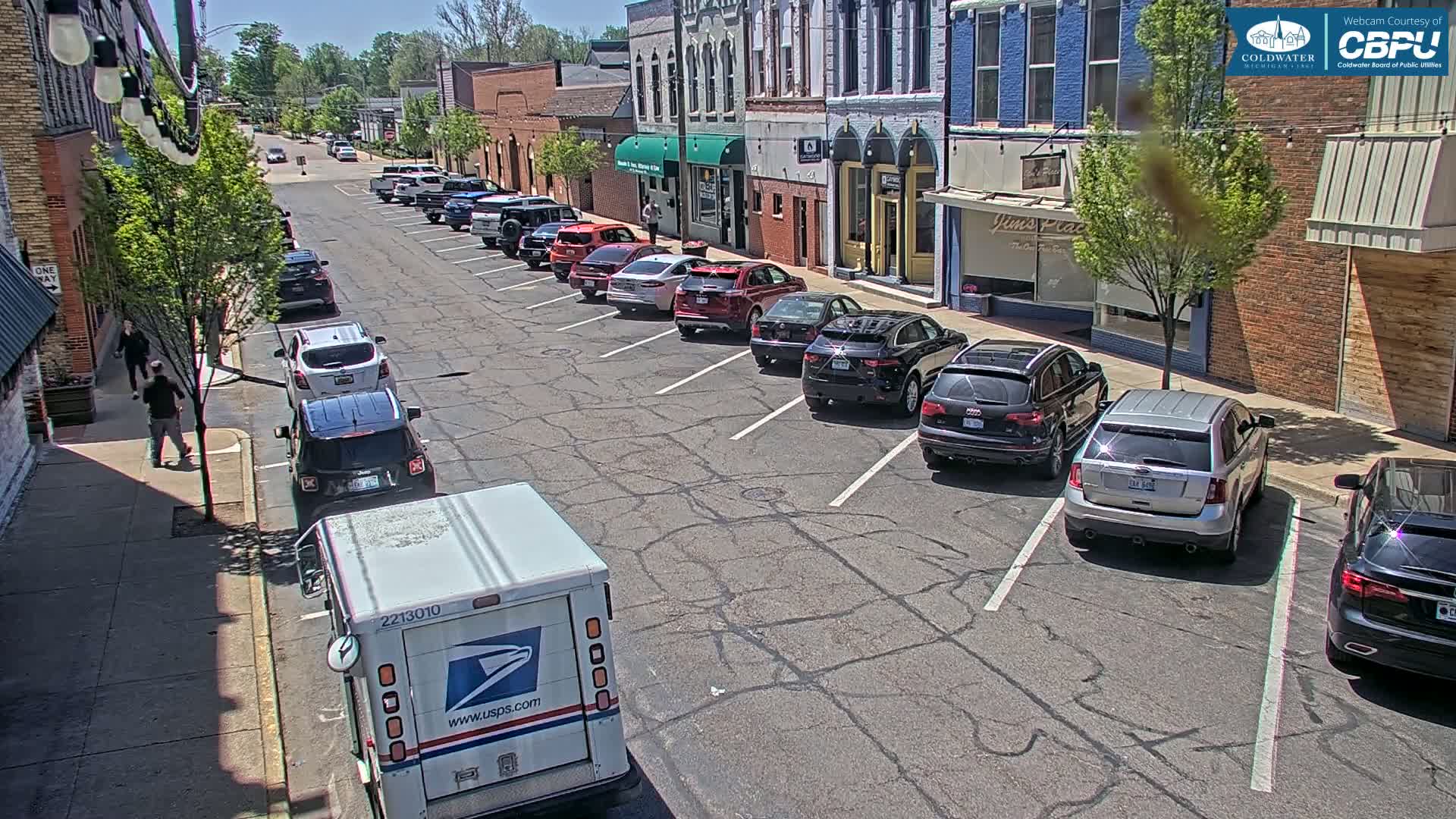 A sunny day shows a street lined with parked cars in front of brick buildings, a postal truck, and two pedestrians walking on the sidewalk.