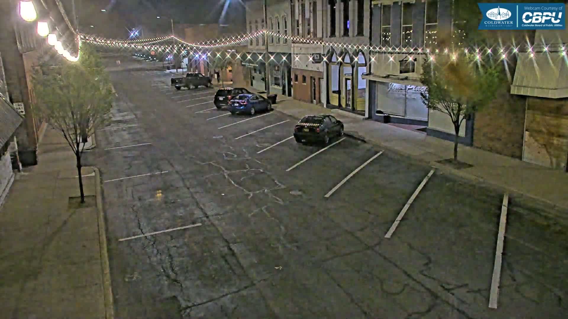 A mostly empty nighttime street lined with parked cars and buildings decorated with string lights.