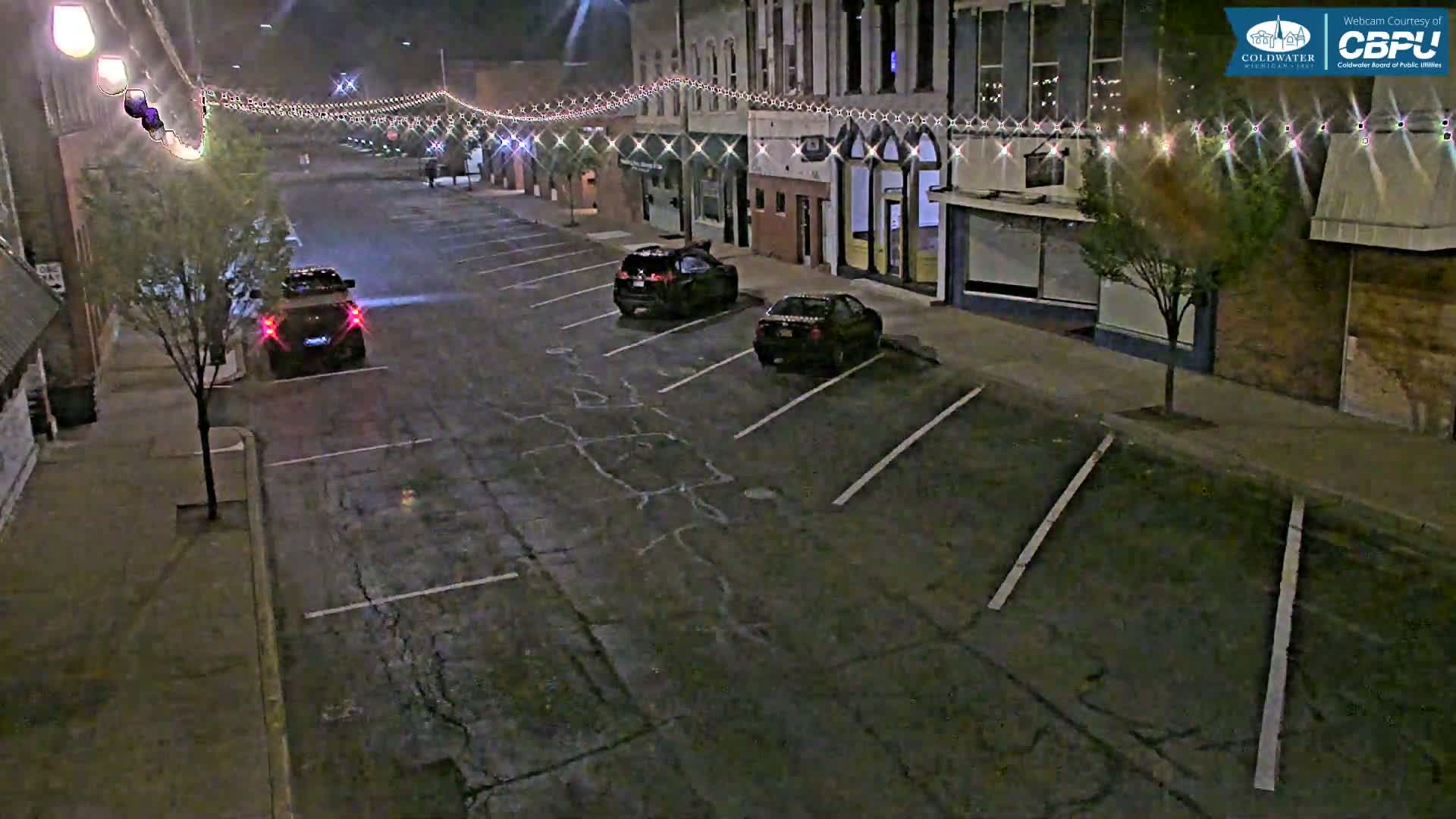 At night, a mostly empty street lined with buildings has string lights overhead and a few cars parked in the lot.
