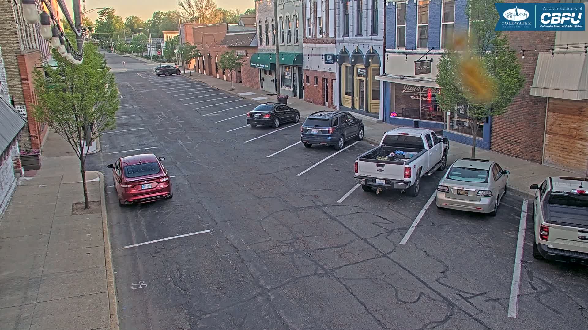 Several cars are parked along a street in front of brick buildings on a partly sunny day.