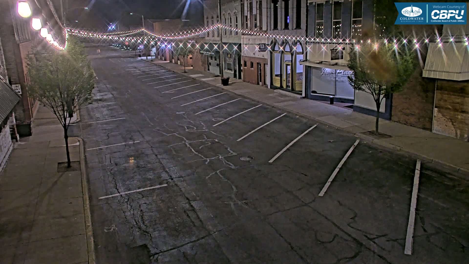 A mostly empty, nighttime street with parked cars, lined with buildings decorated with string lights.