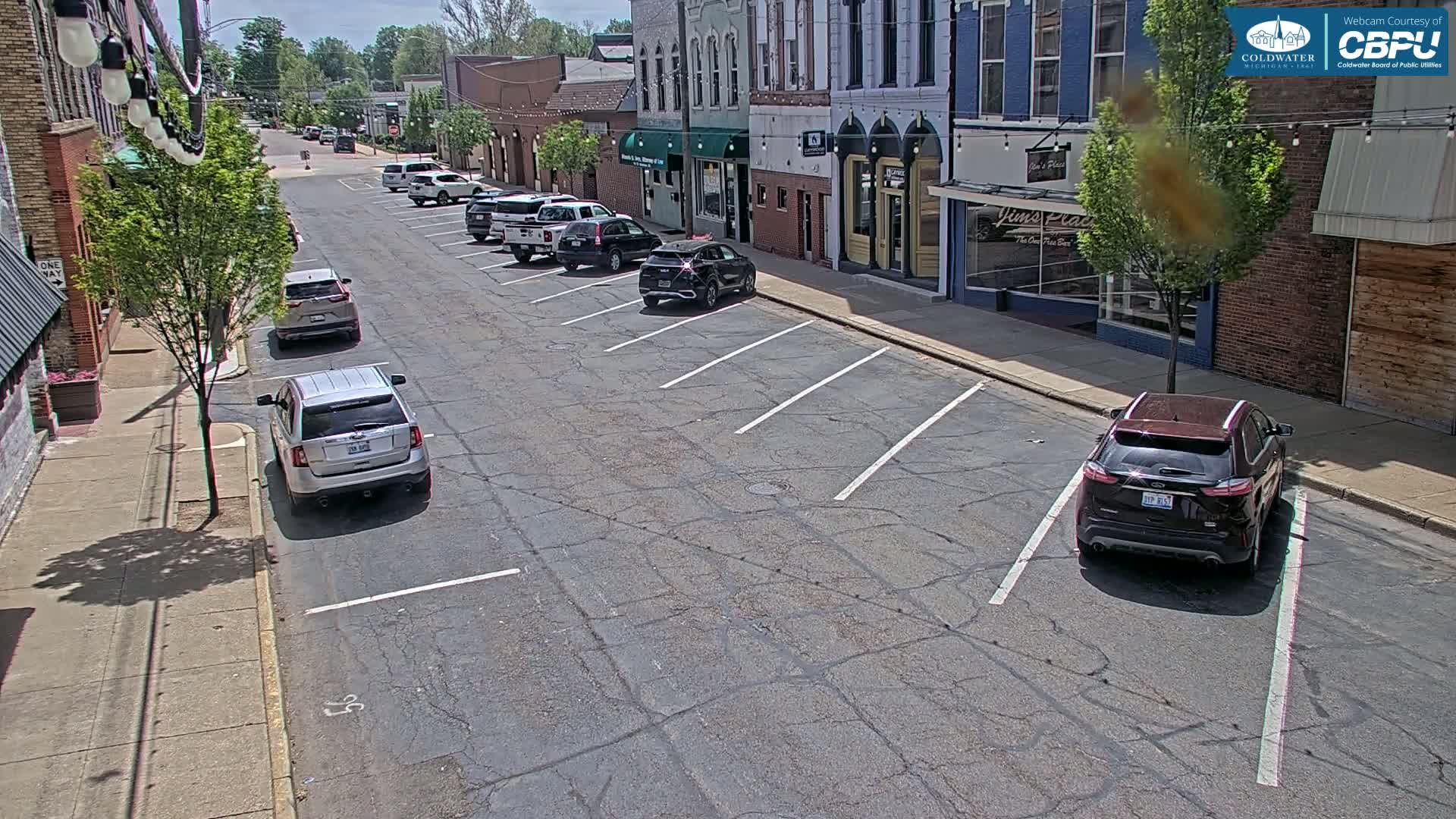 A sunny street scene shows several cars parked along the curb and in parking spaces in front of brick buildings.