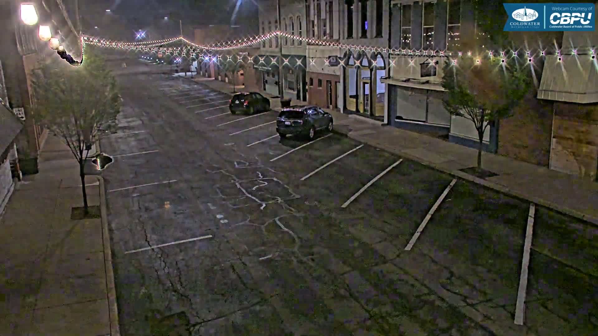 A nighttime street scene shows a nearly empty parking lot in front of brick buildings adorned with strings of lights.