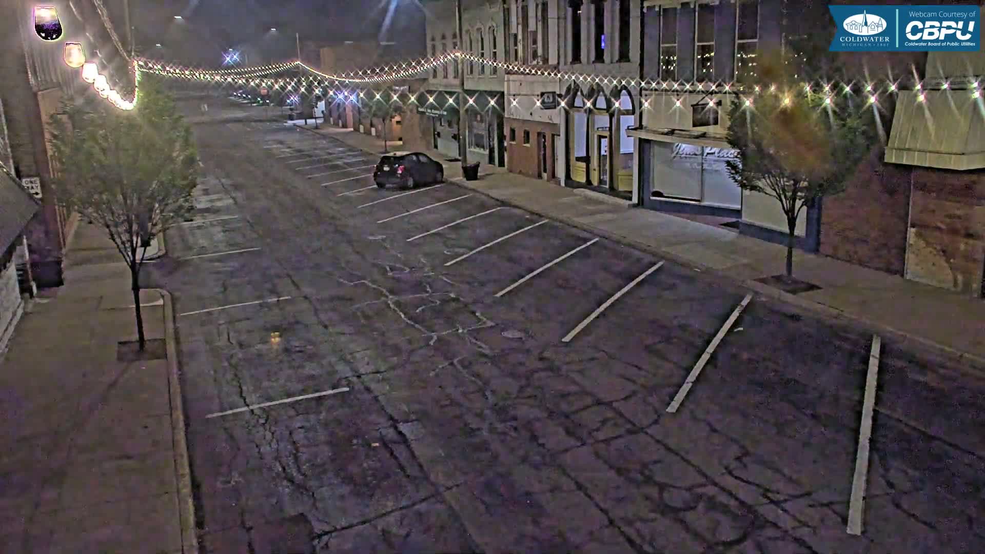 A nearly empty street at night, lined with buildings and string lights, is lightly trafficked by a single car parked at the side.