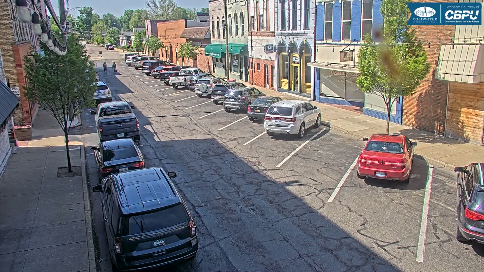 Cars are parked along a sunny street lined with brick buildings on a partly cloudy day.