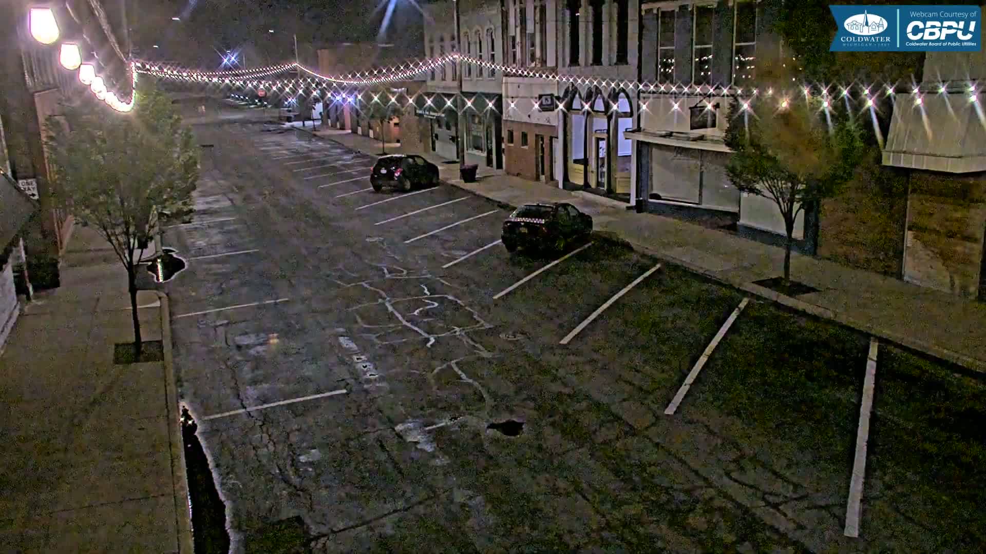 A nighttime street scene shows a mostly empty parking lot in front of brick buildings illuminated by string lights.