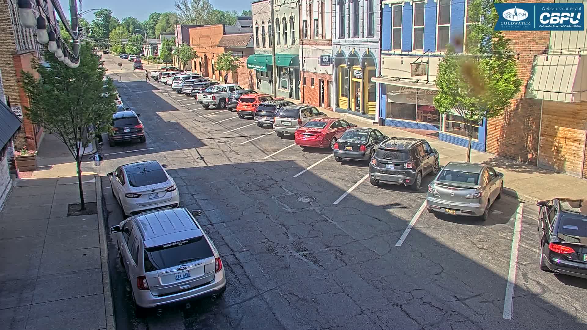 Cars are parked along the side of a street in front of brick buildings on a sunny day.
