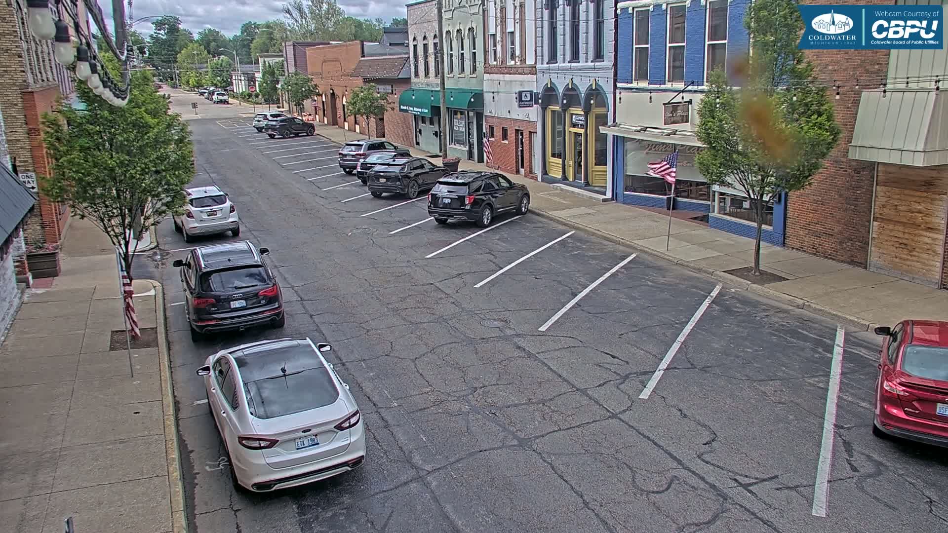 A street with parked cars lines the sides of brick buildings under a partly cloudy sky.