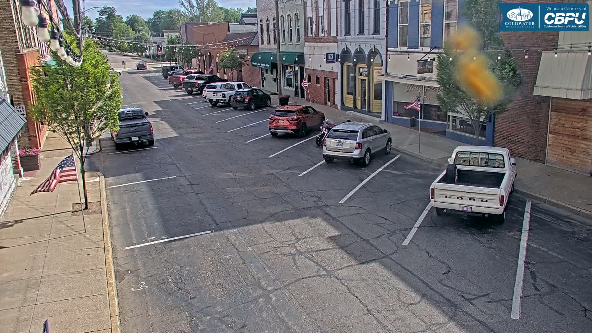 Several cars and trucks are parked along a sunny street in front of brick buildings on a partly sunny day.