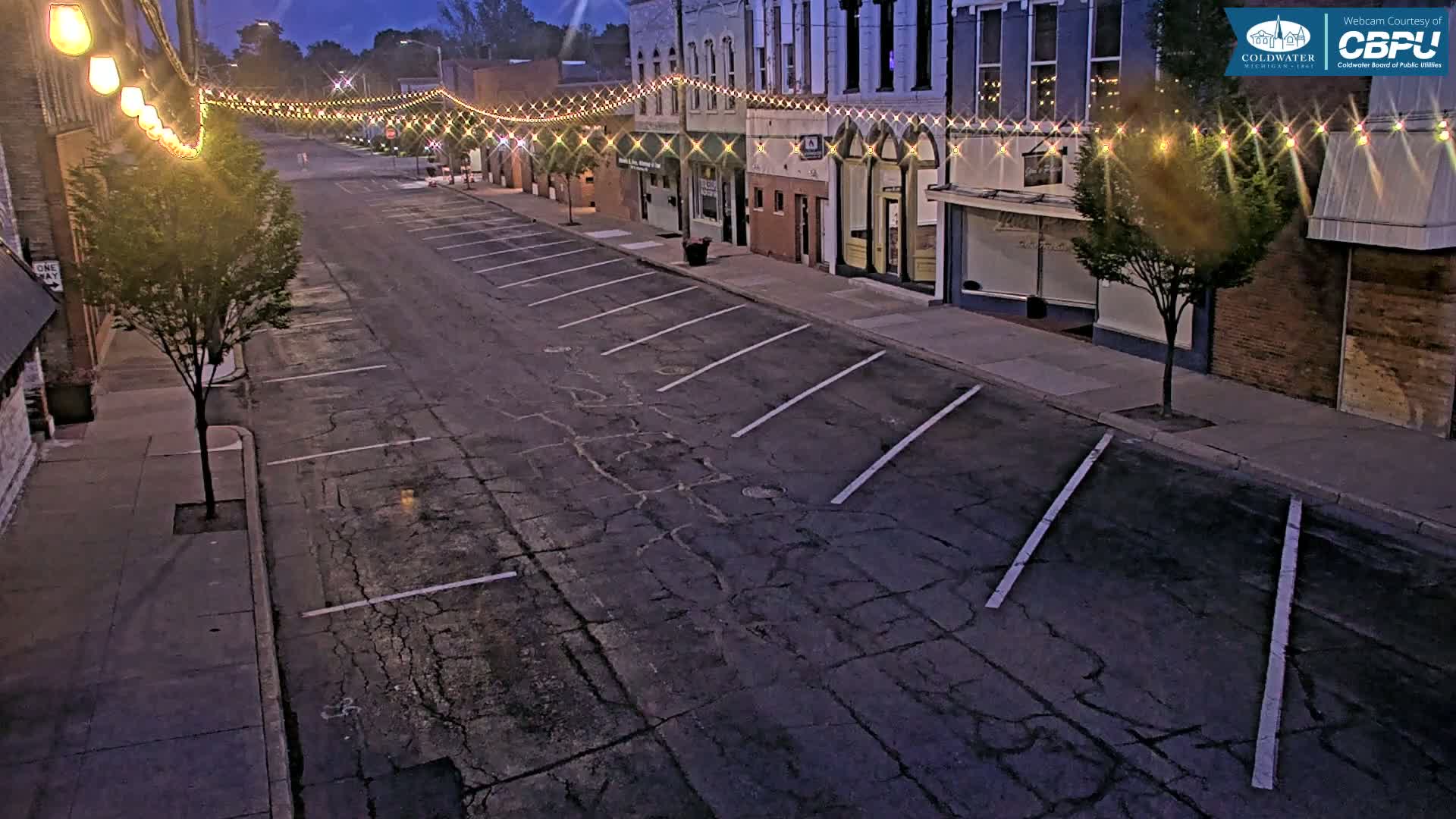 A mostly empty street at dusk lined with buildings decorated with string lights, showing a cracked asphalt parking lot.