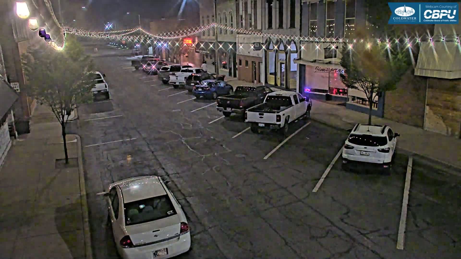 A nighttime street scene shows several cars parked along the curb of a street lined with buildings decorated with string lights under a dark, possibly overcast sky.