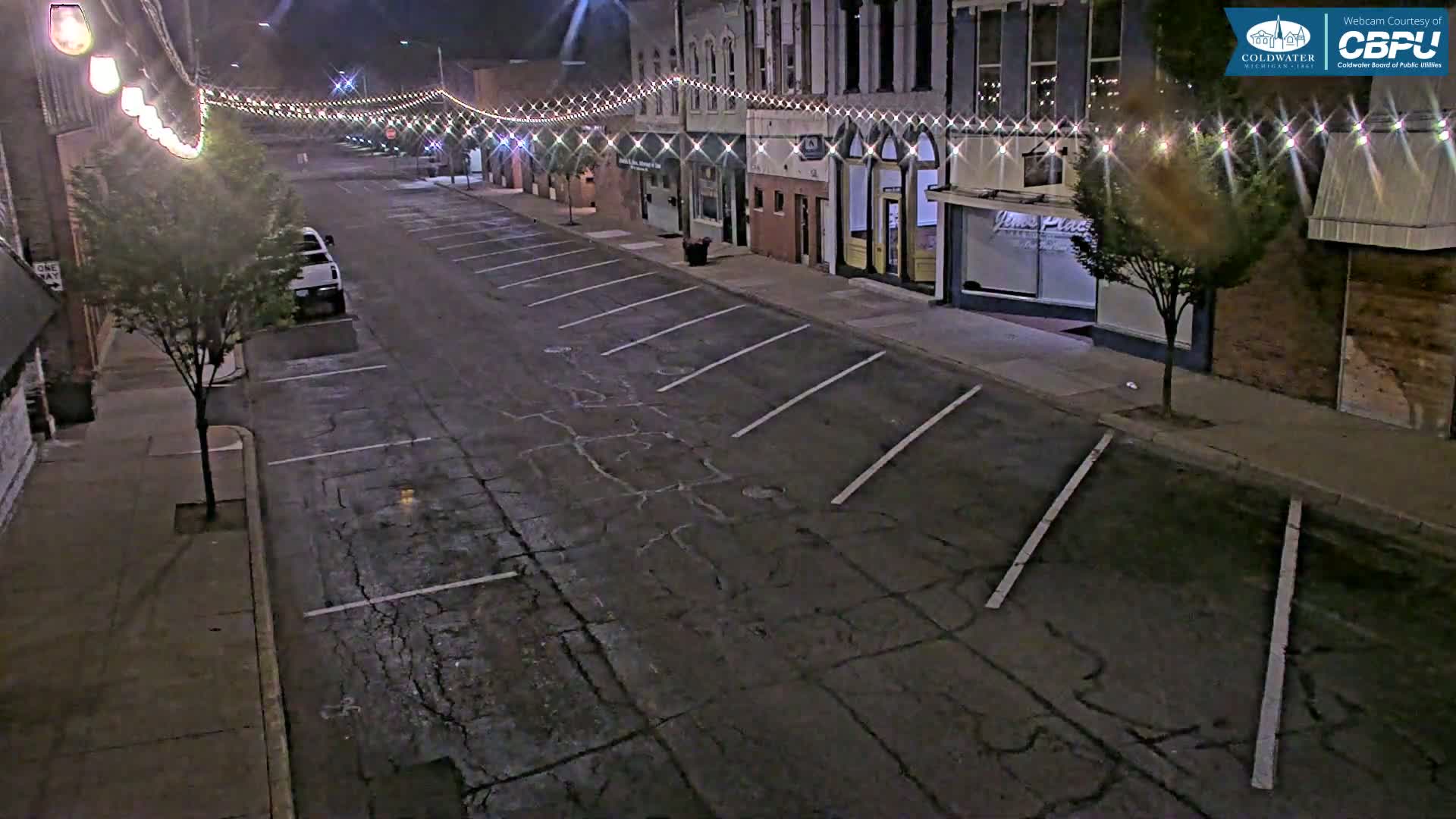 A mostly empty street at night, lined with parked cars and buildings decorated with string lights, under a dark sky.