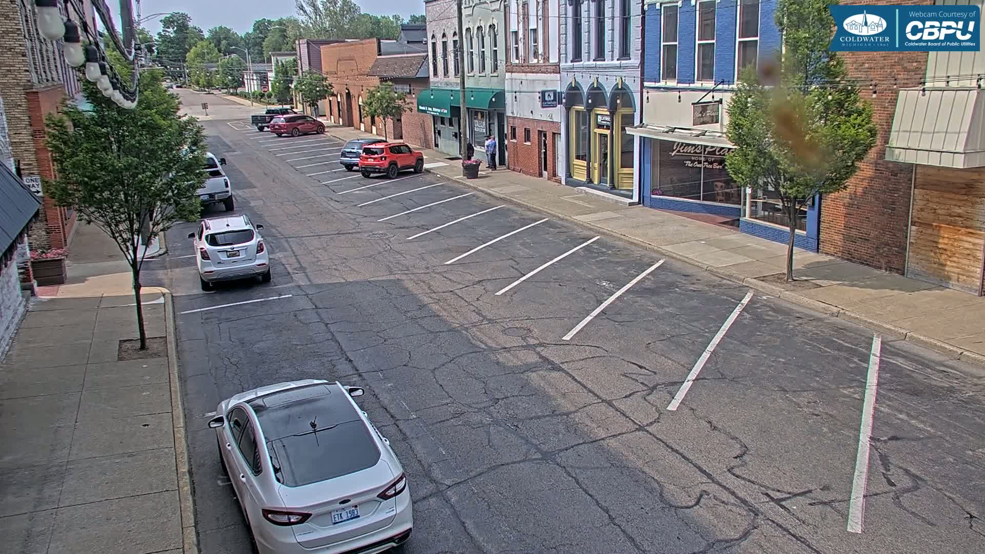 A mostly sunny street scene shows a row of parked cars along the street in front of brick buildings on a partly sunny day.