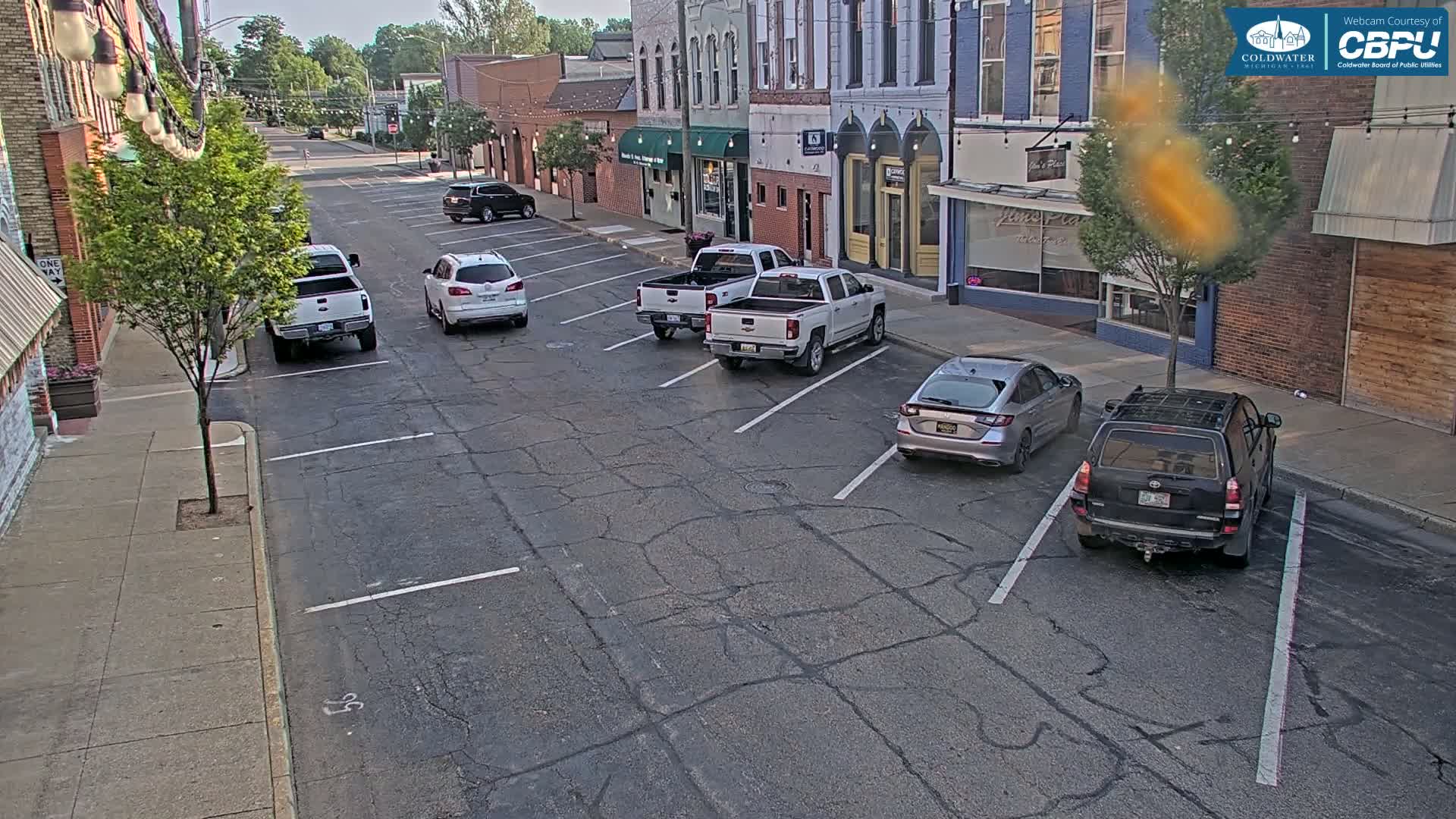 Several vehicles are parked along a street in front of brick buildings on a partly sunny day.