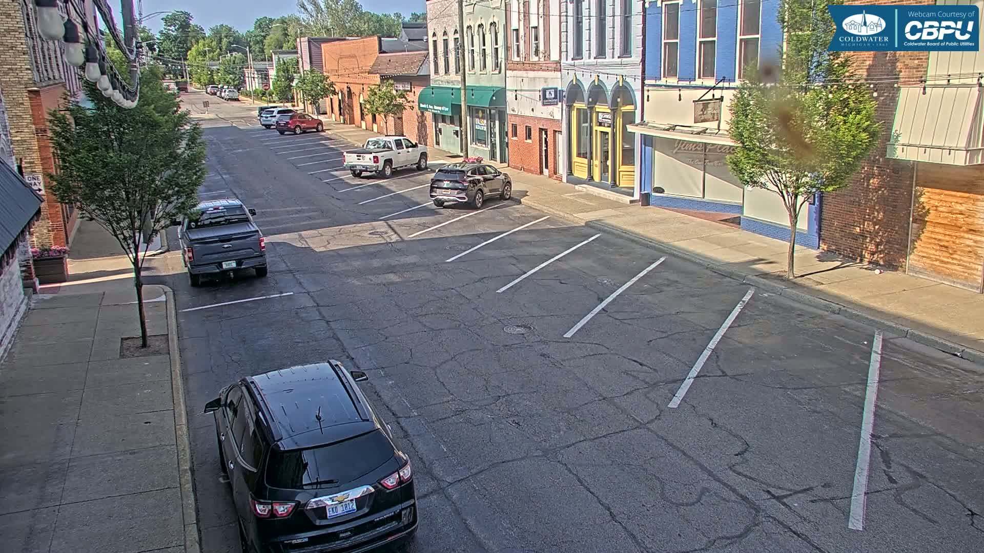 A sunny street scene shows several parked cars in front of brick buildings on a mostly empty street.