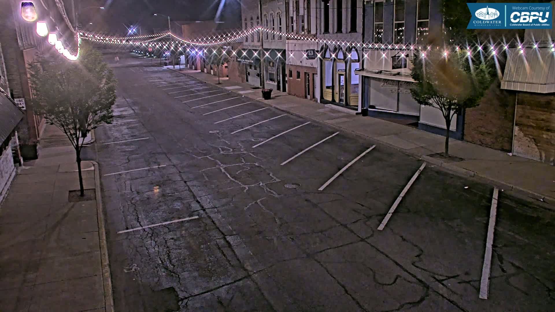 A mostly empty street at night, lined with buildings and illuminated by string lights, shows parking spaces along the side of the road.