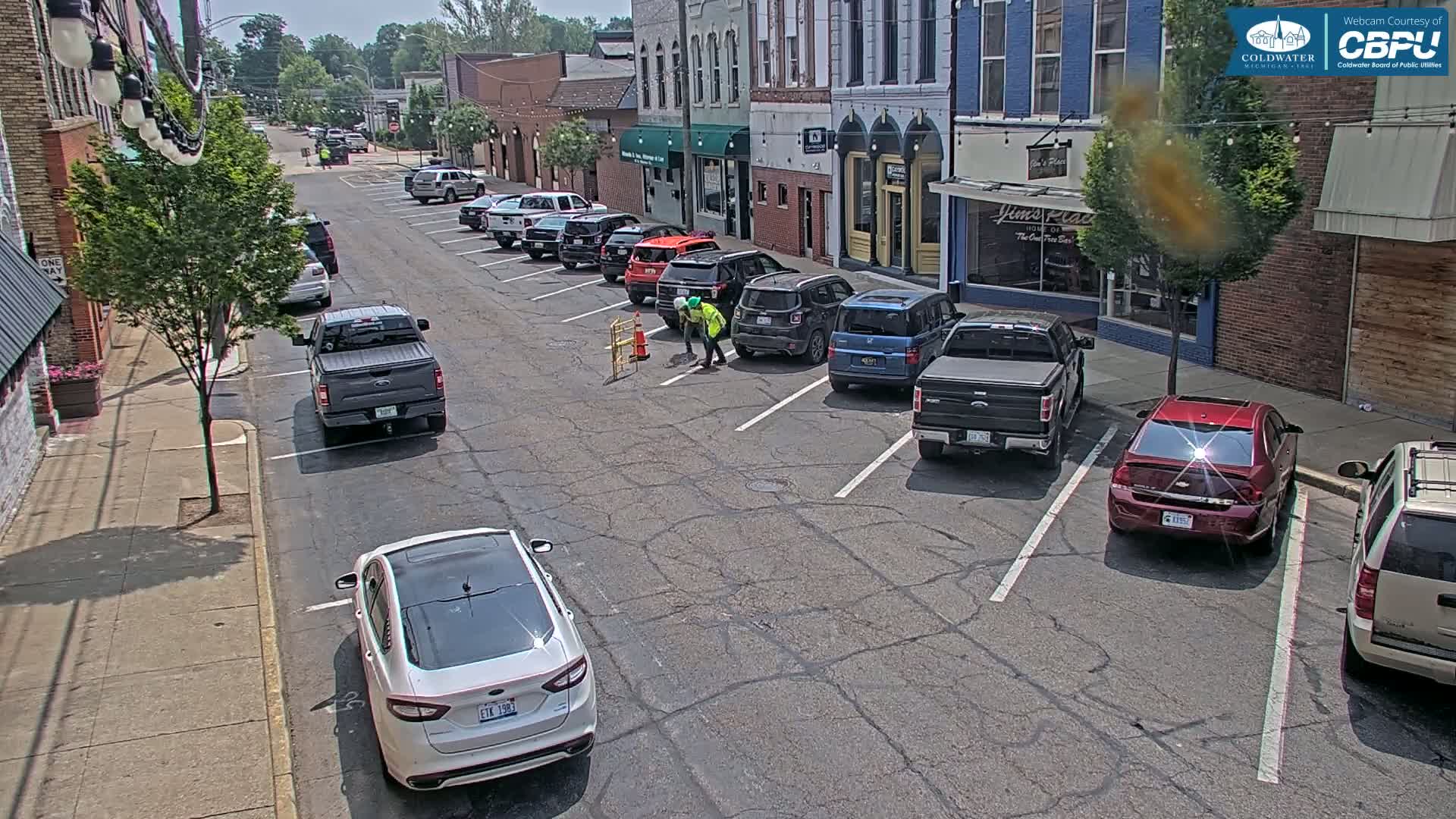 On a sunny day, two workers are repairing a street in a town's parking lot lined with cars parked along the curb.