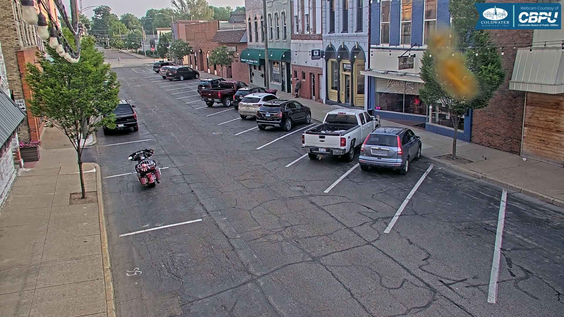 A mostly empty street with parked cars lines the sides of brick buildings on a partly sunny day.