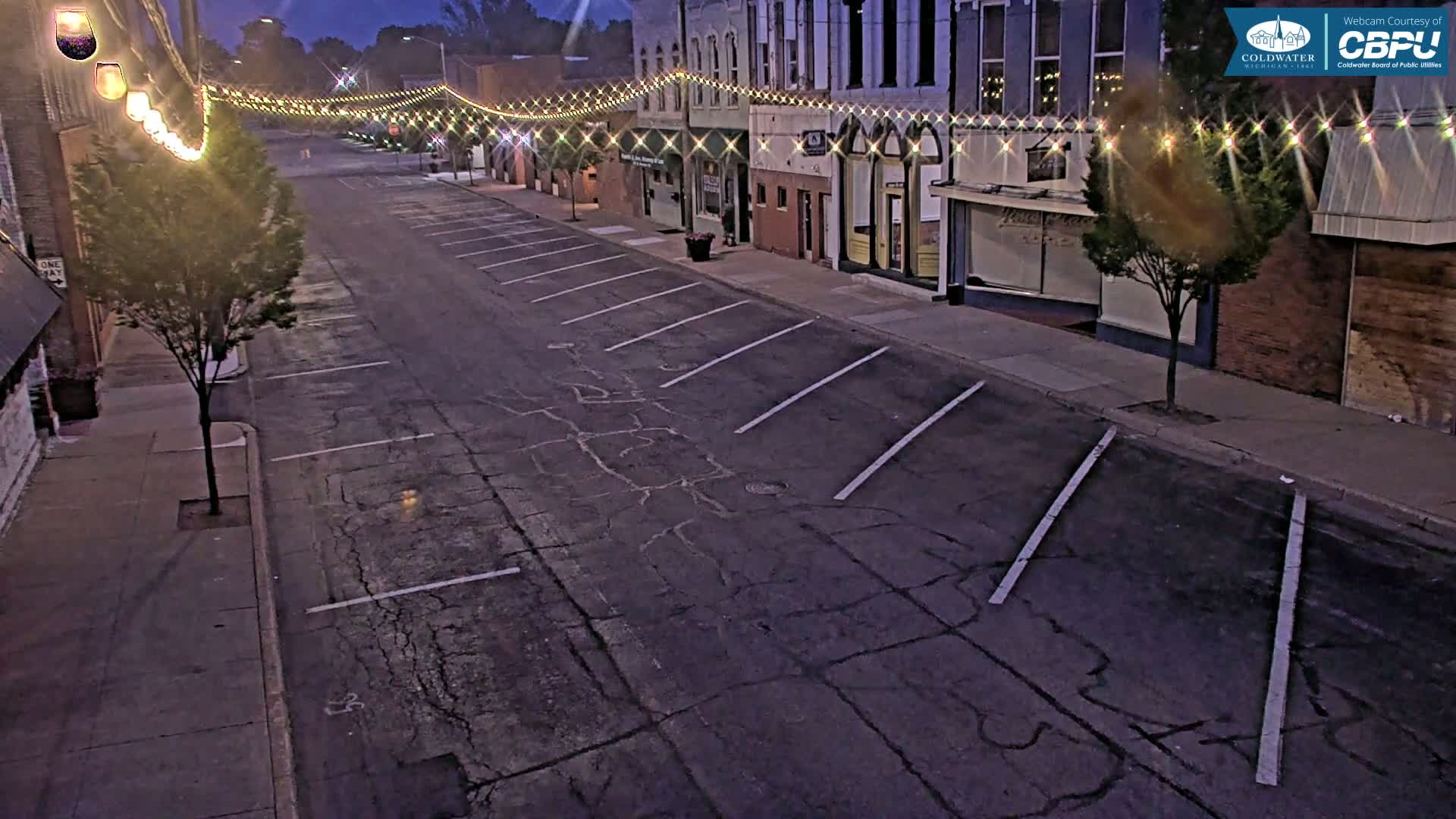 A mostly empty street at dusk, lined with buildings and string lights, shows some parking spaces and a cracked asphalt road.