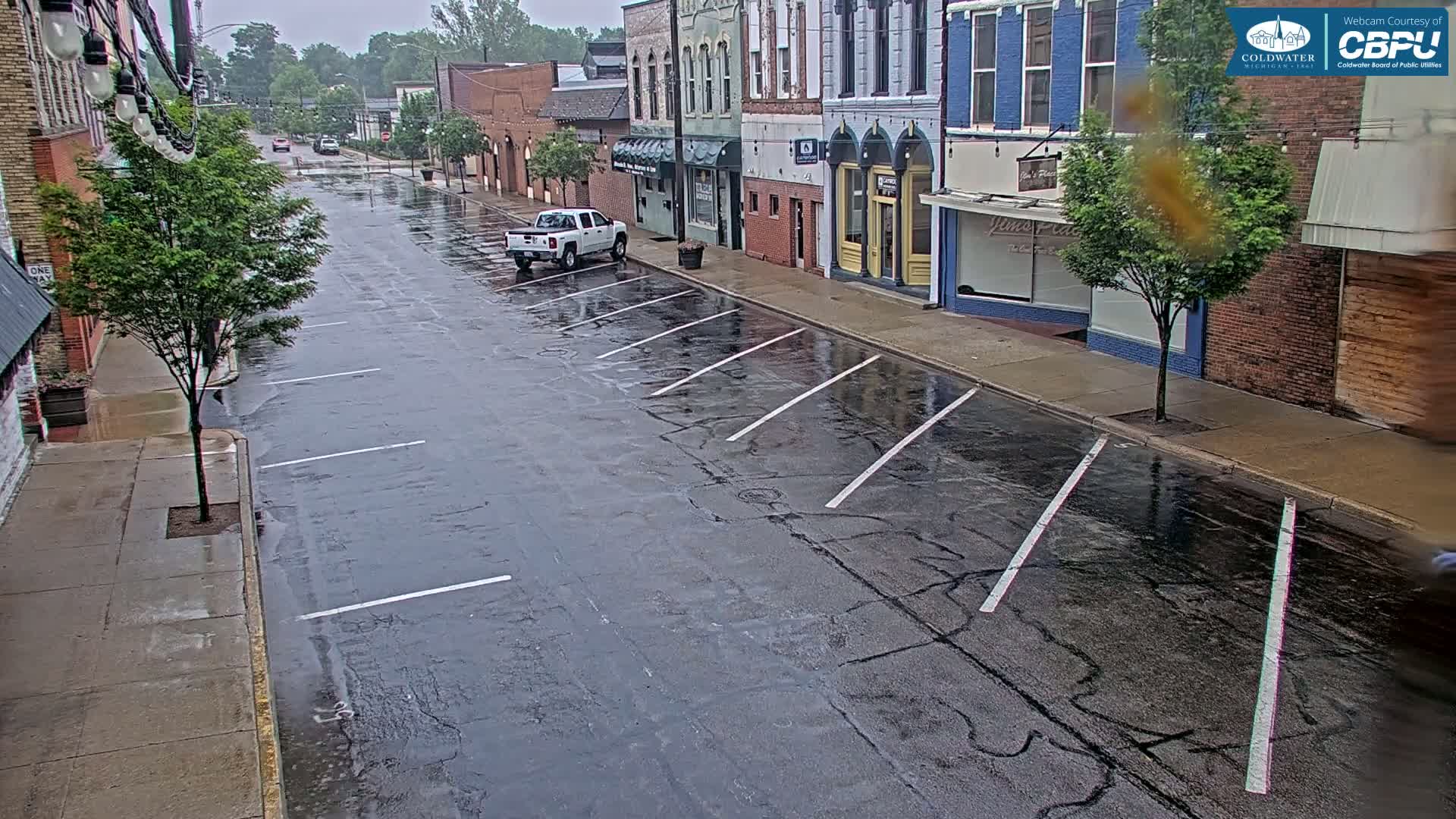 A wet street with parked cars and shops lines either side under an overcast sky.