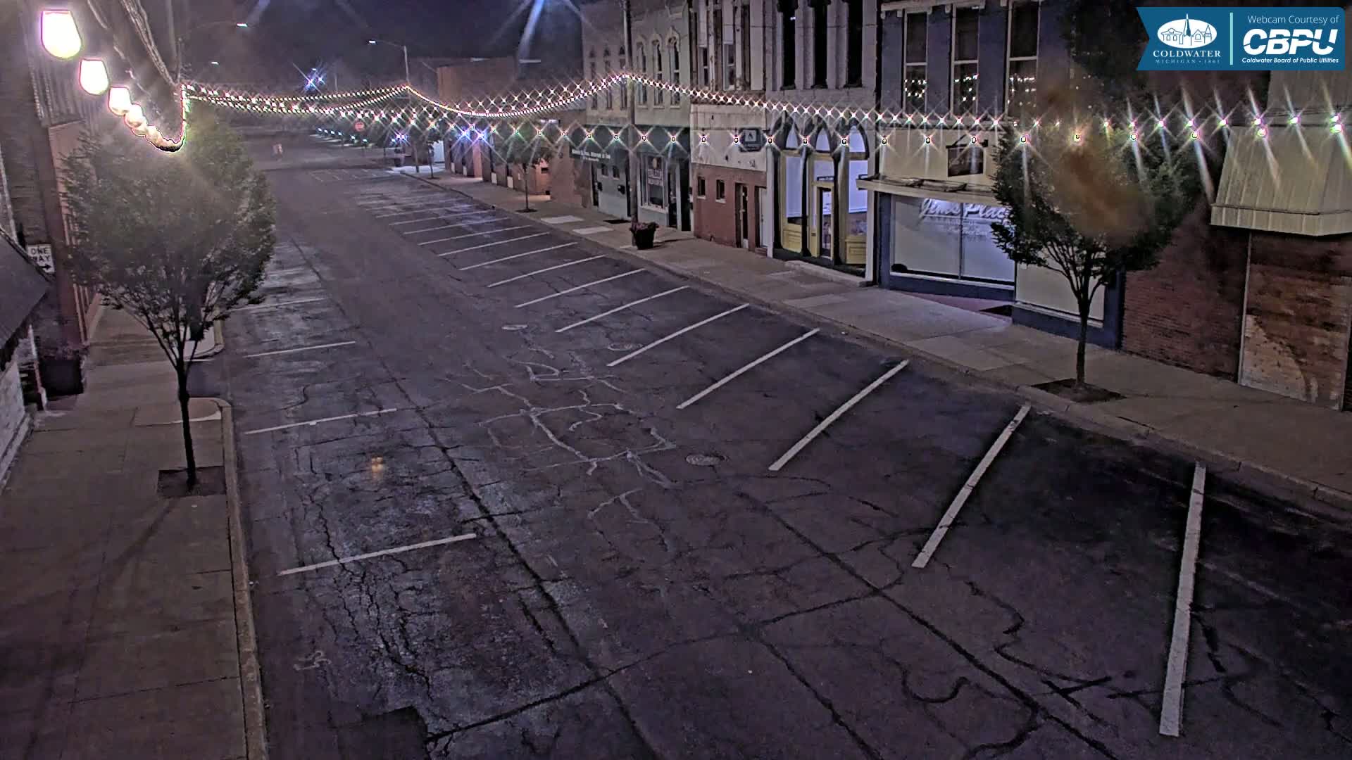 An empty street at night, lined with brick buildings decorated with string lights, shows cracked asphalt parking spaces and small trees.