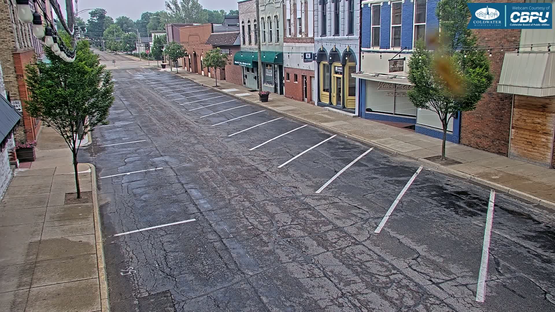 A mostly empty street with parallel parking spaces on a cloudy day shows several brick buildings with various storefronts.
