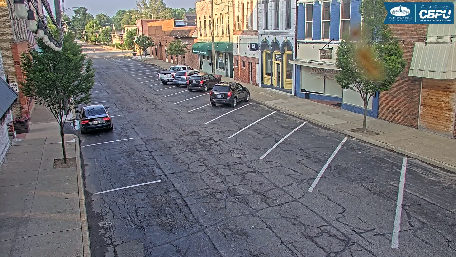 A mostly empty street with several cars parked along the curb in front of brick buildings on a partly sunny day.