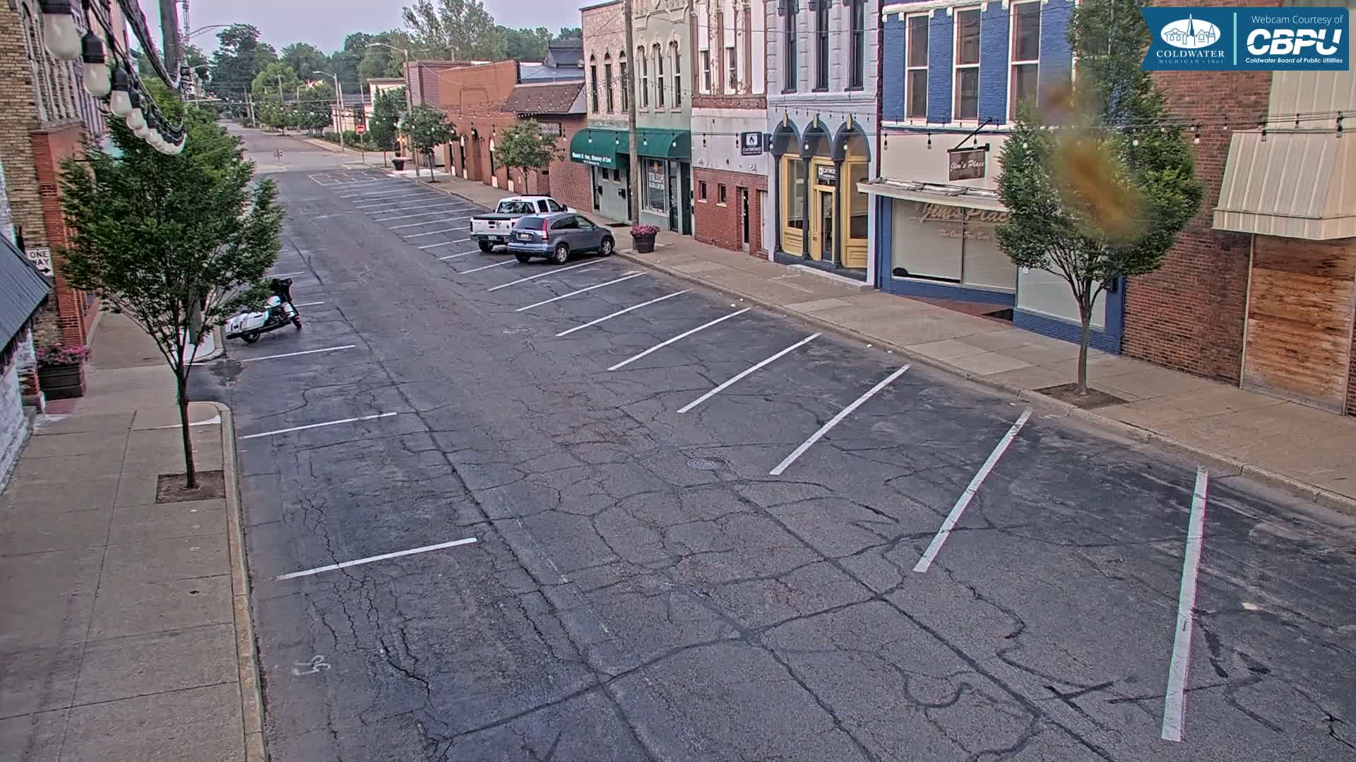 A mostly empty street with parked cars and a motorcycle in front of brick buildings on a partly cloudy day.
