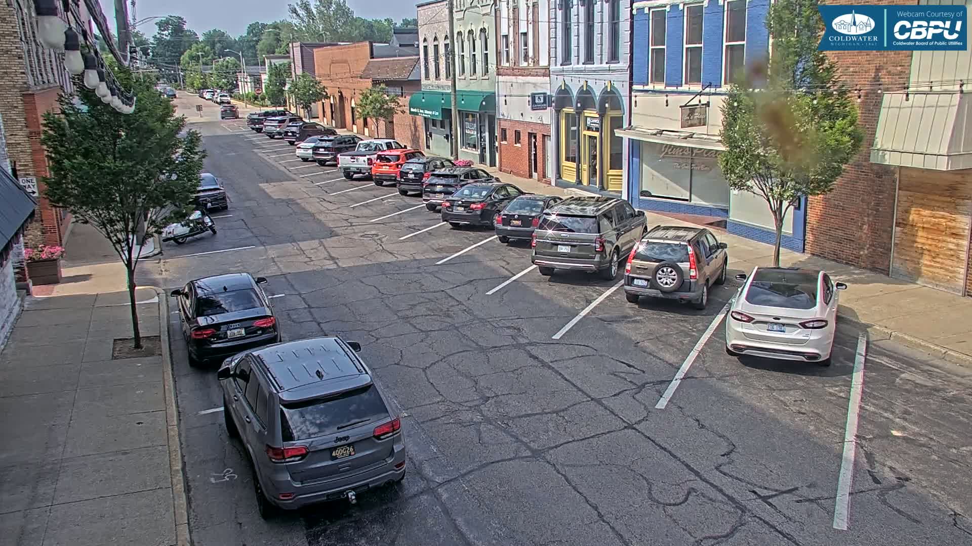 A sunny street scene shows several cars parked along the side of the road in front of brick buildings.