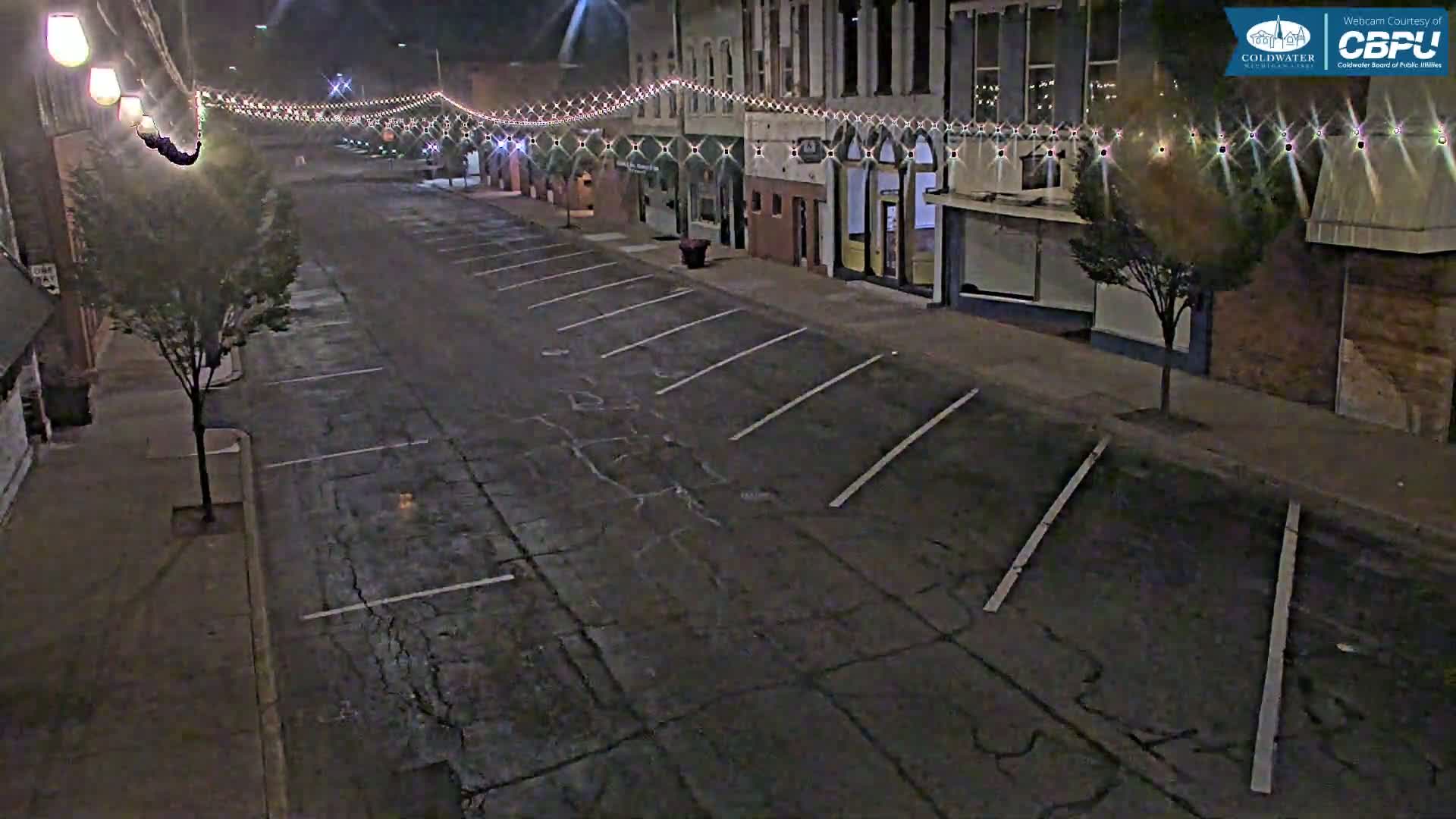 An empty street at night, lined with buildings decorated with string lights, is shown under dark conditions.